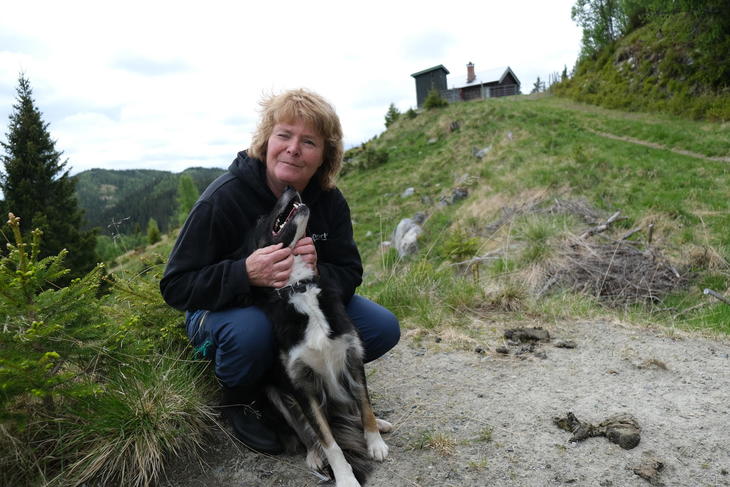 Grethe Ringdal sitter på ei gresstue og koser med en av gjeterhundene. I bakgrunn ser vi en seterstøl.