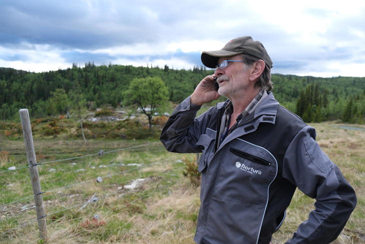 John Bergerud, kledd i Nortura-klær med tydelig logo, er på fjellet og snakker i telefonen. Han står ved et gjerde.
